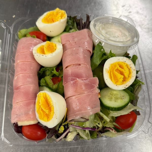 A chef salad with ham, hard-boiled eggs, tomatoes, cucumbers, and mixed greens in a plastic container.