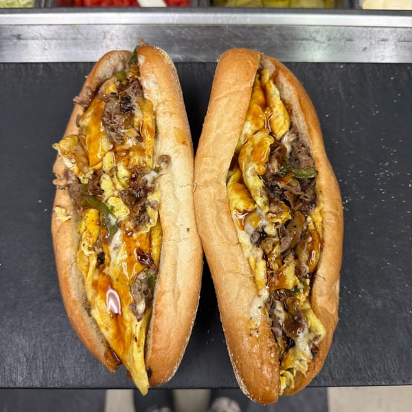 Two hearty Sabrin Pizza steak and egg subs with melted cheese and sauce on a dark surface.