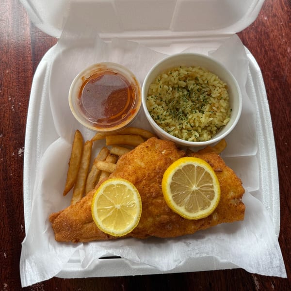 Fried fish with lemon slices, french fries, rice, and sauce in a takeout container.