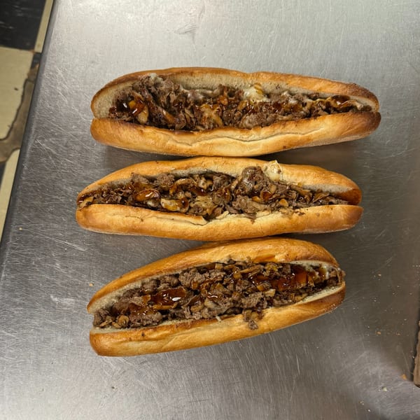Three Sabrin Pizza cheesesteak sandwiches with meat, cheese, and sauce on a metal surface.